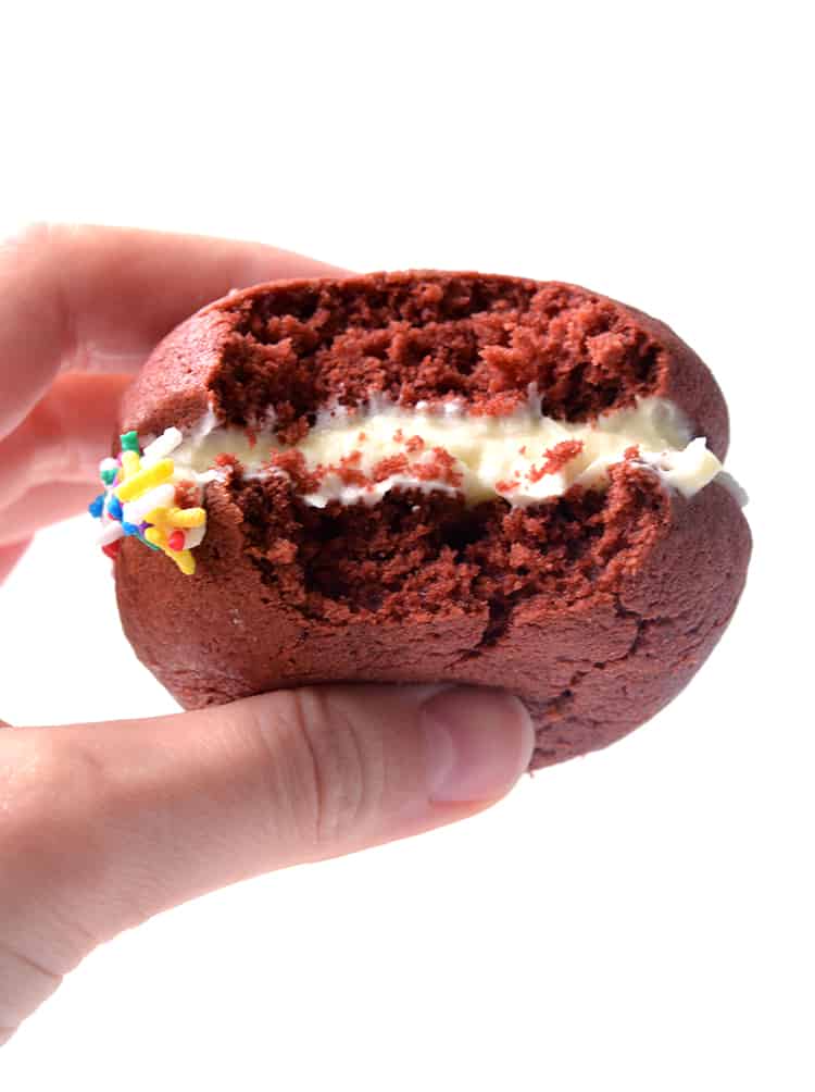 A hand holding a red velvet whoopie pie with a bite taken out of it