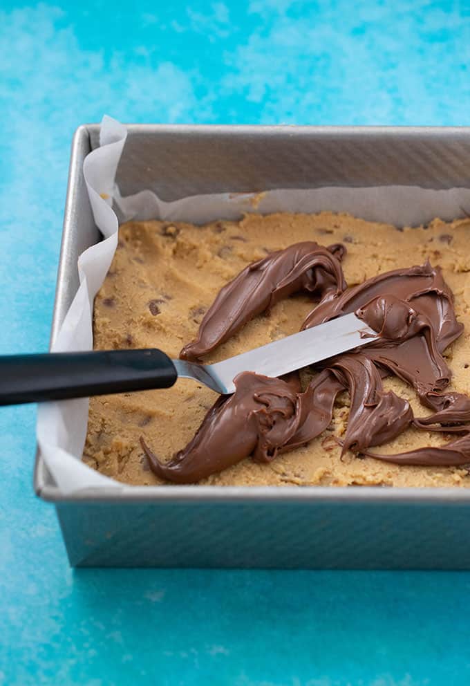 A baking tray filled with chocolate chip cookie dough and topped with spoonfuls of Nutella
