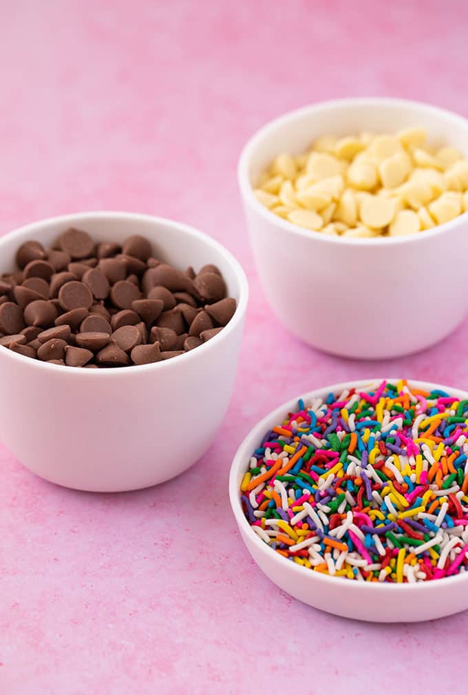 Bowls filled with chocolate chips and sprinkles on a pink background