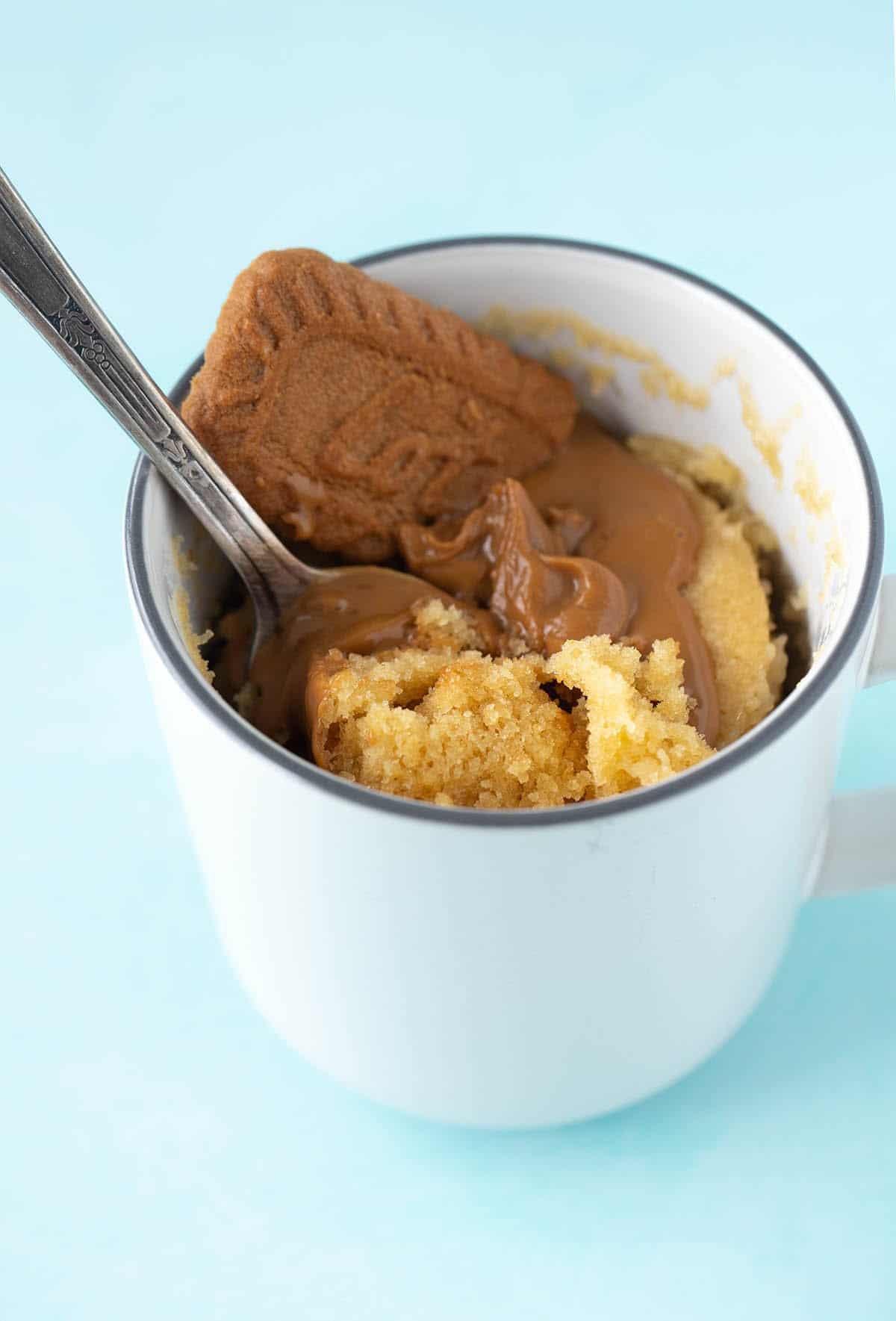 A spoon diving into a Biscoff Mug Cake on a blue background.