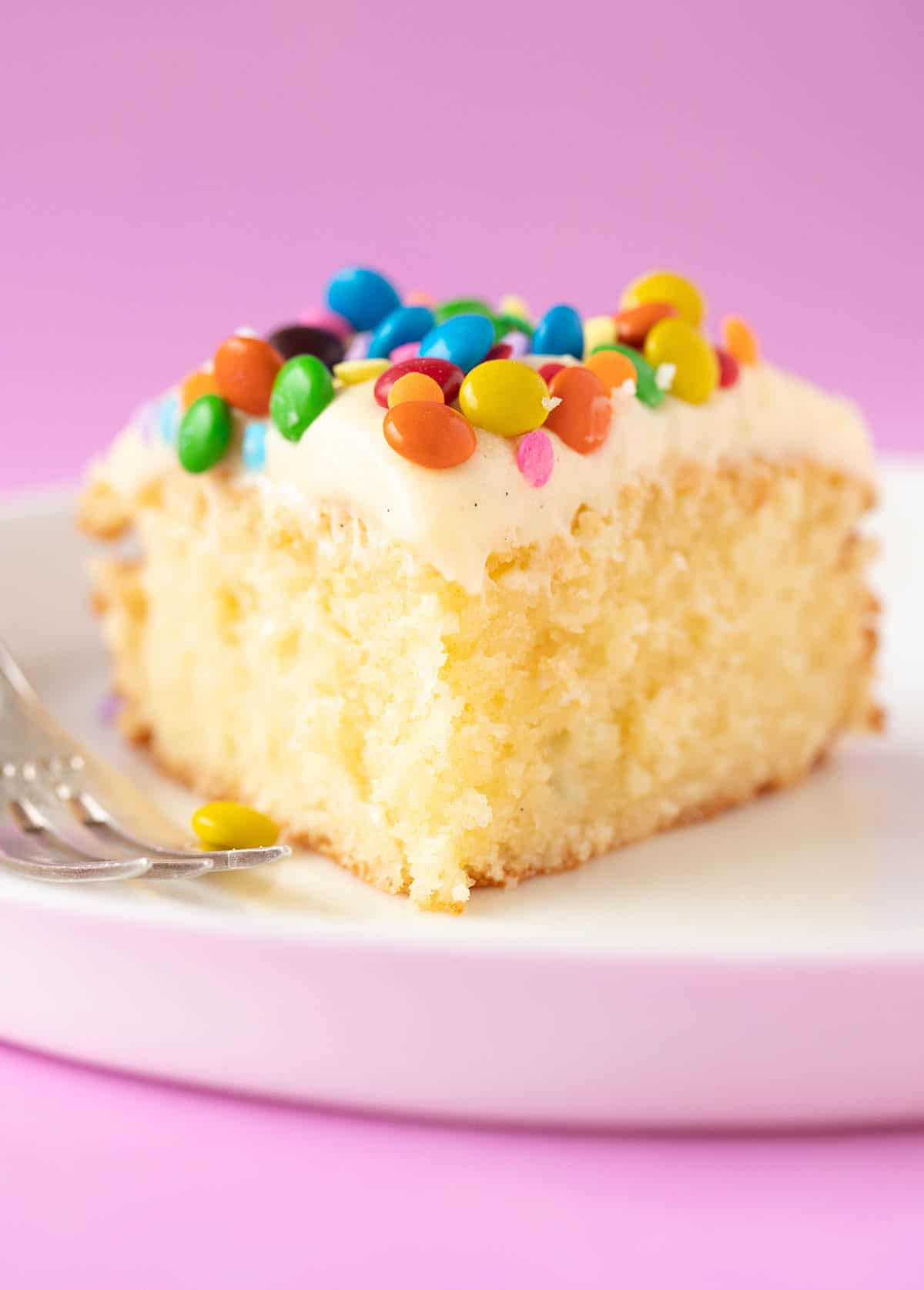 Close up view of a big slice of Vanilla Cake topped with vanilla frosting and M&M's.