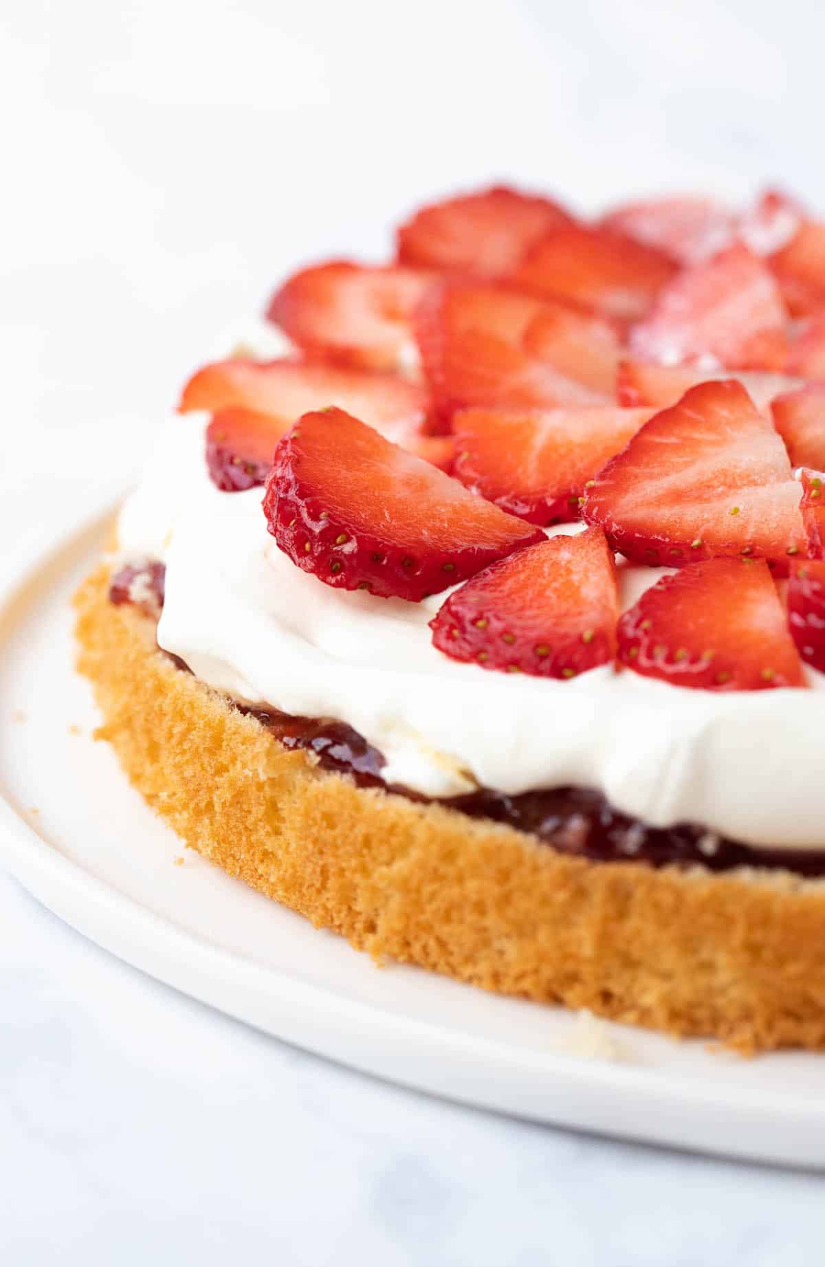 A sponge cake topped with jam, cream and strawberries on a white plate.