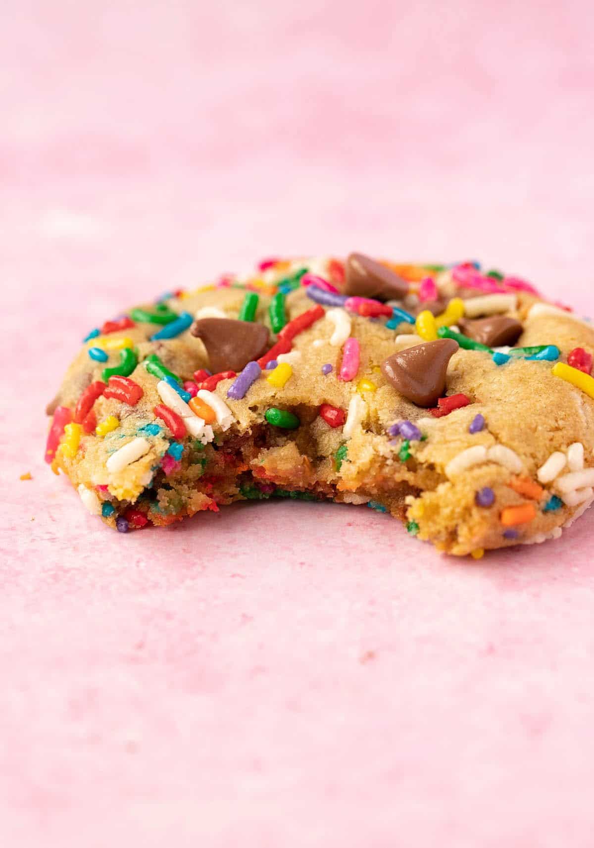  A Funfetti Chocolate Chip Cookie with a bite taken out it on a pink background.