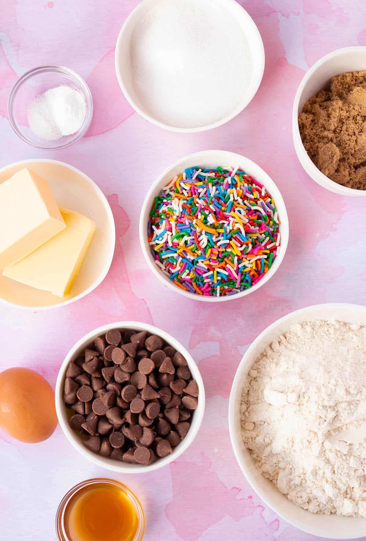 All the ingredients needed to make Funfetti Cookies laid out on a pink background. 