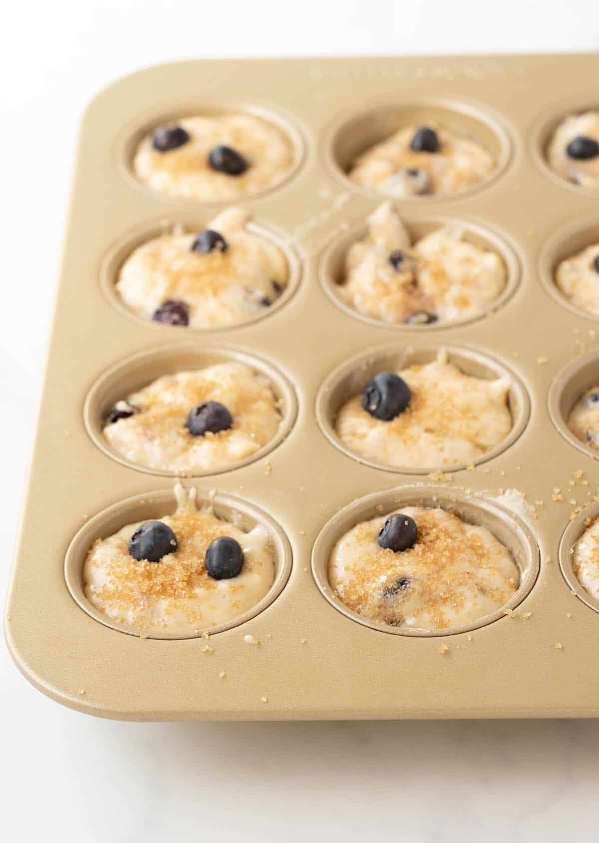 A gold muffin pan filled with blueberry muffin batter.