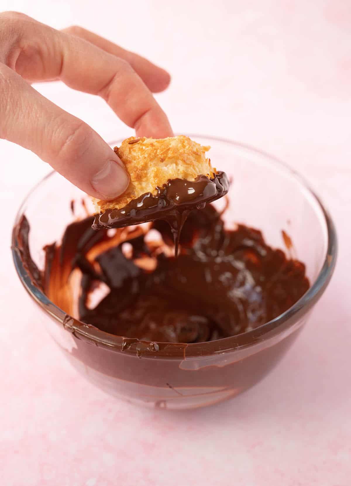A hand dipping a Coconut Macaroon in a bowlful of melted chocolate.