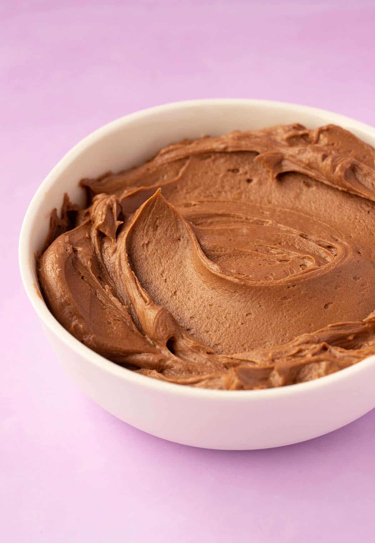 A bowl of creamy chocolate frosting on a purple backdrop. 