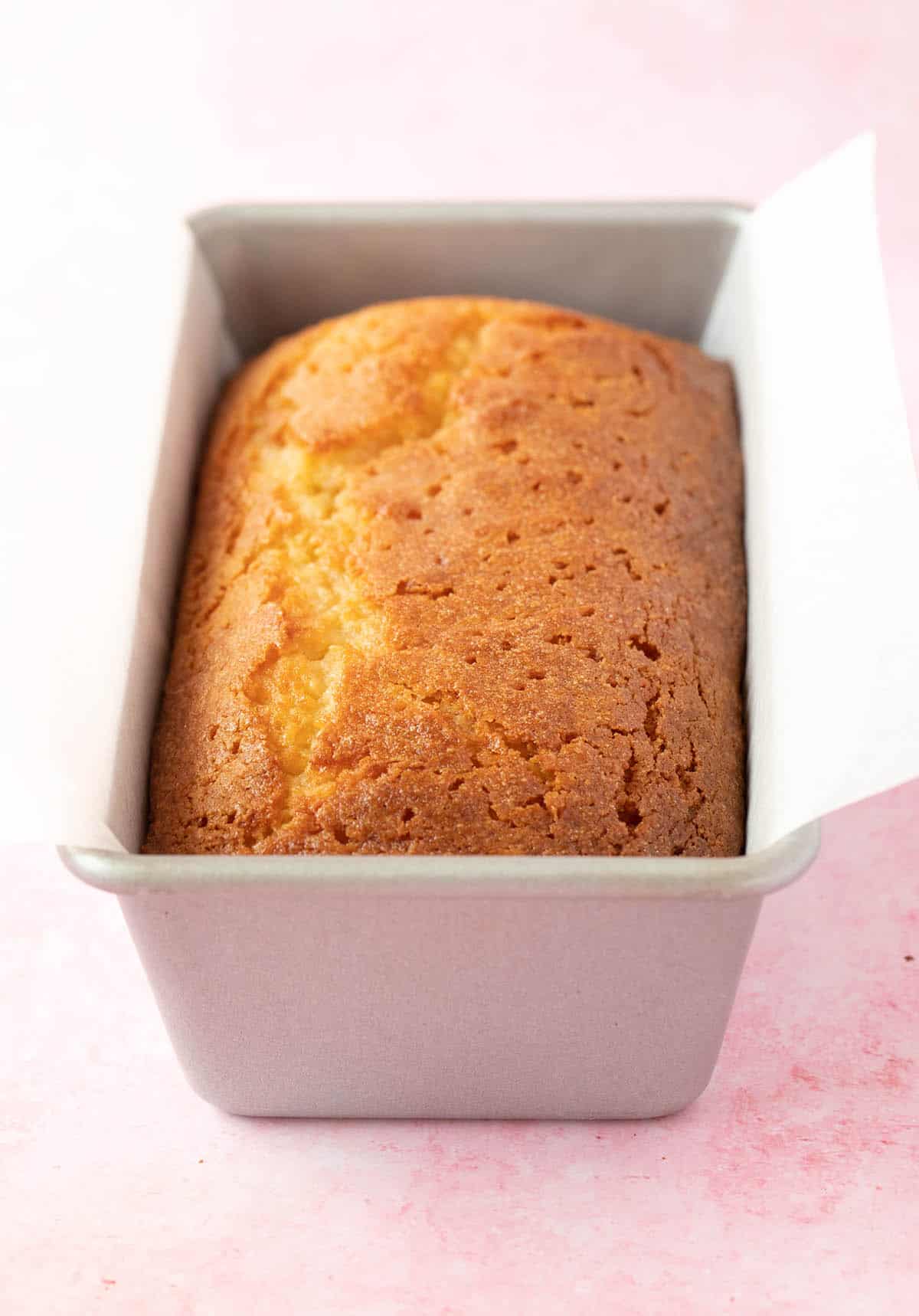 A Lemon Loaf cake sitting in a cake pan on a pink background.