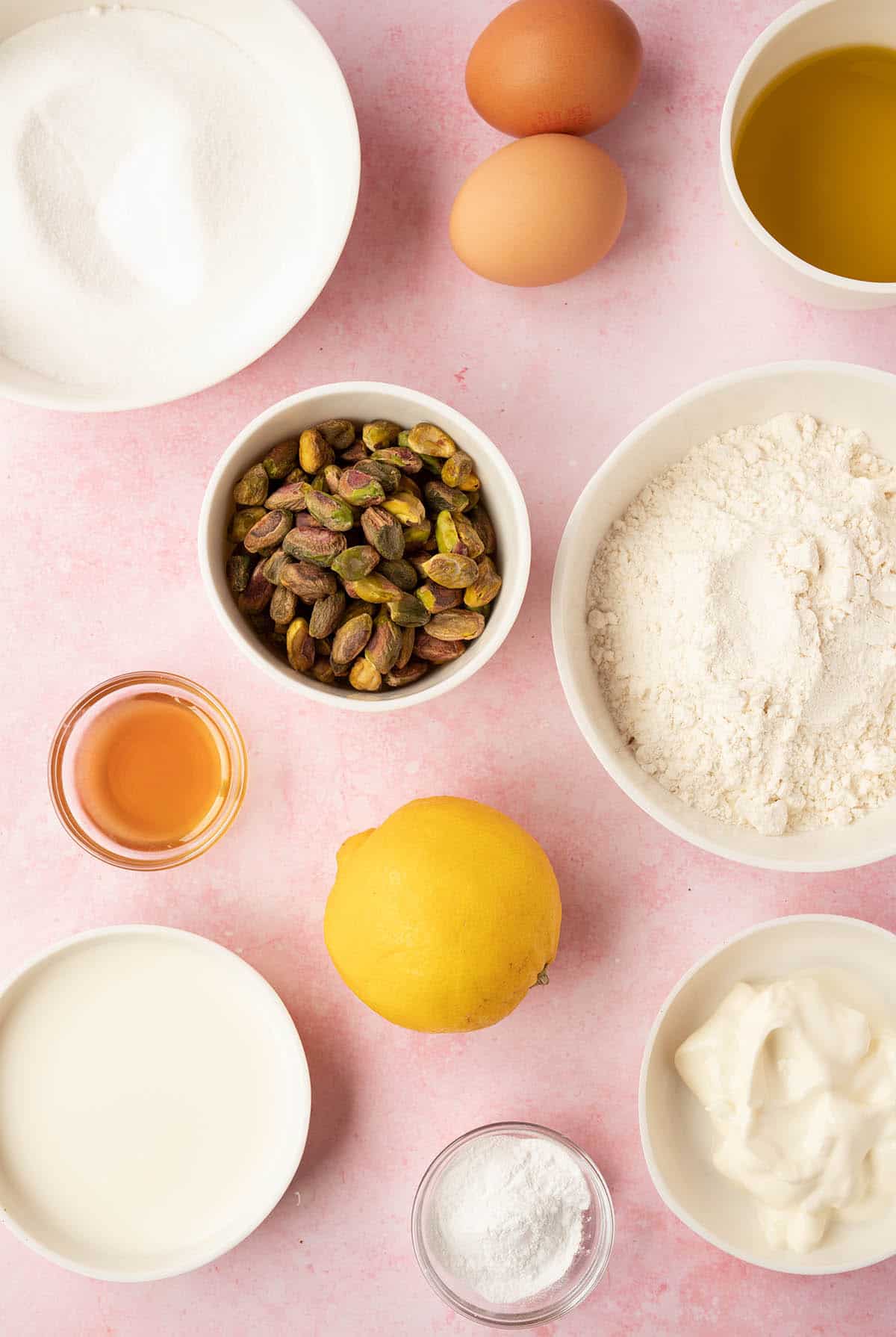 All the ingredients you need to make a Pistachio Cake from scratch sitting on a pink background.