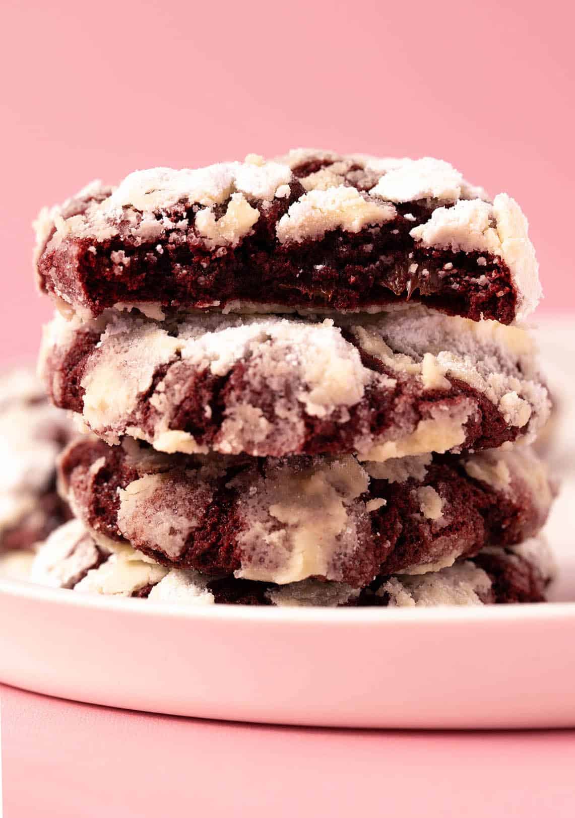A stack of Red Velvet Crinkle Cookies with a bite taken out of it sitting on a white plate.