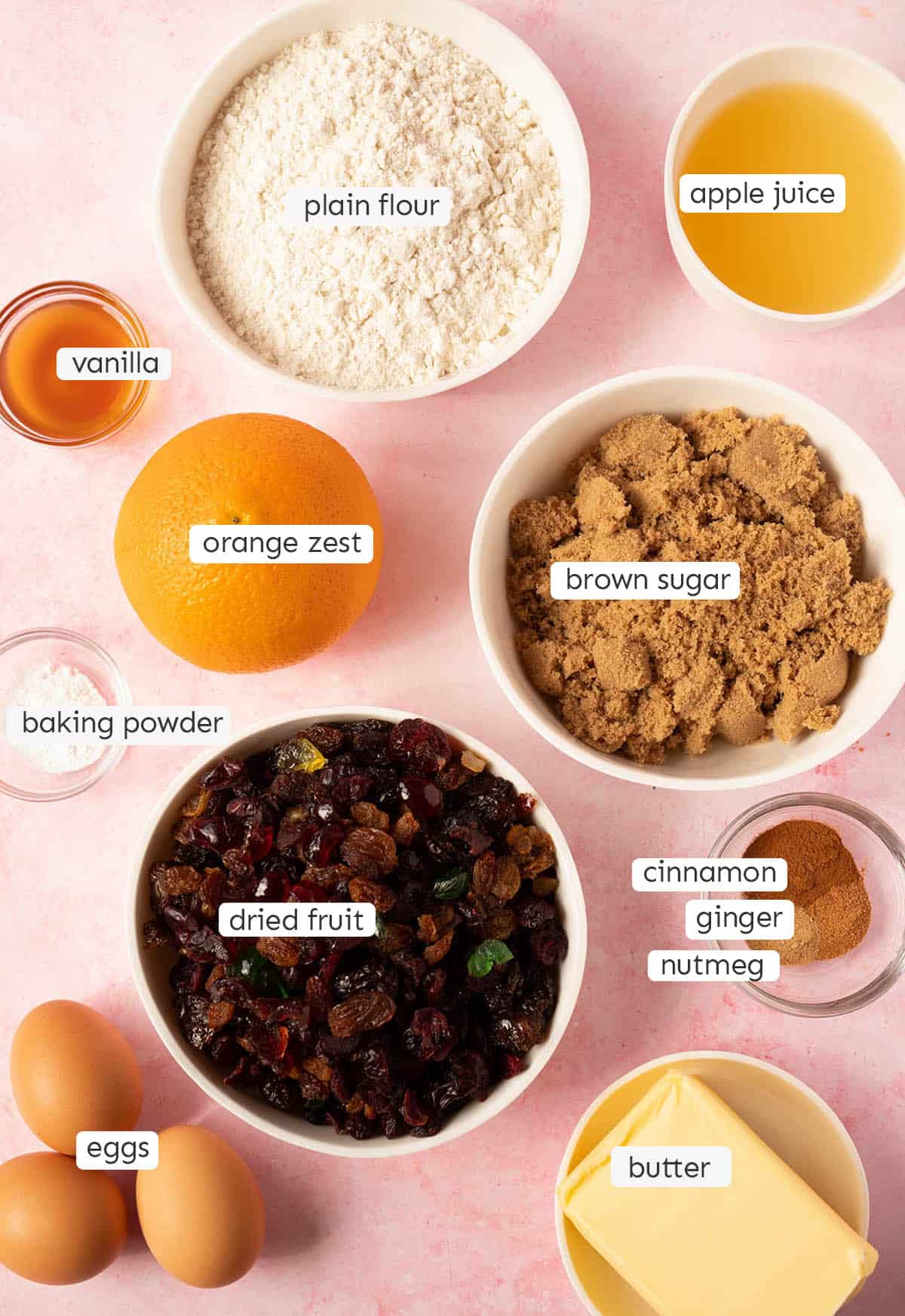 All the ingredients needed to make fruit cake from scratch laid out on a pink backdrop.