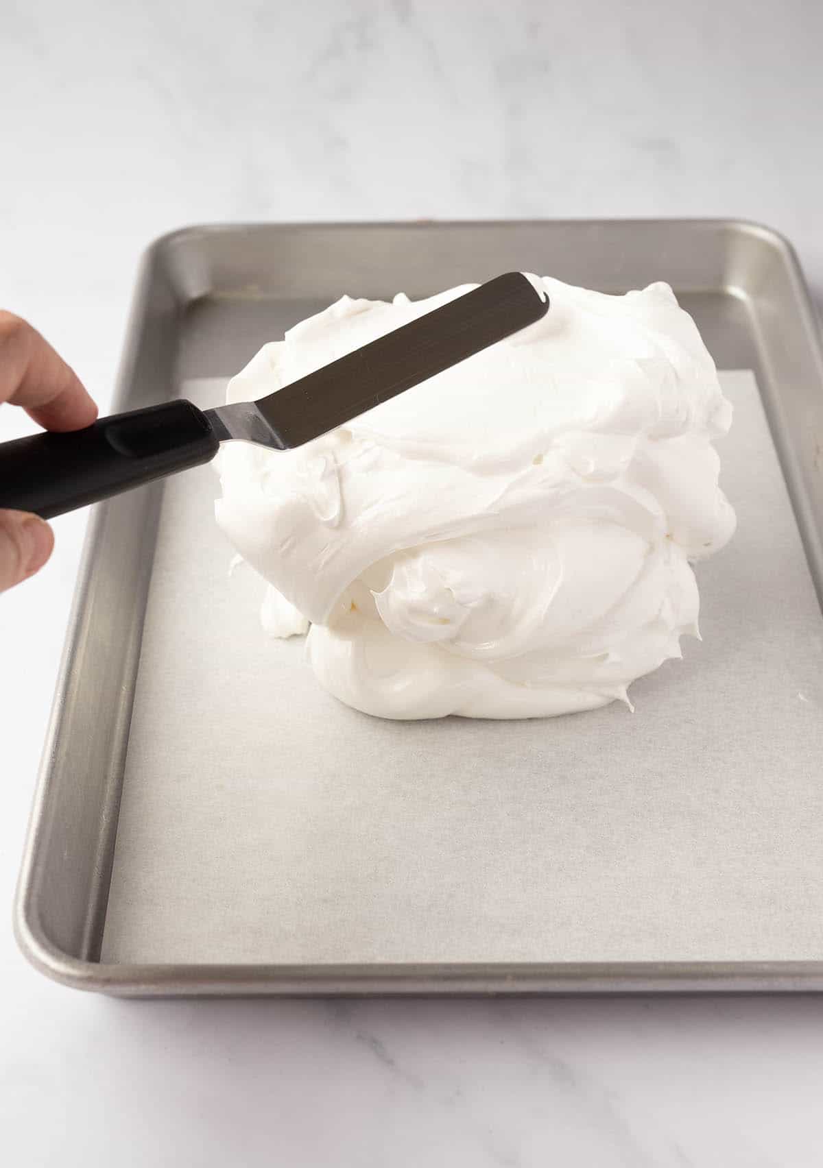 A baking tray topped with meringue with a hand showing how to smooth it out.