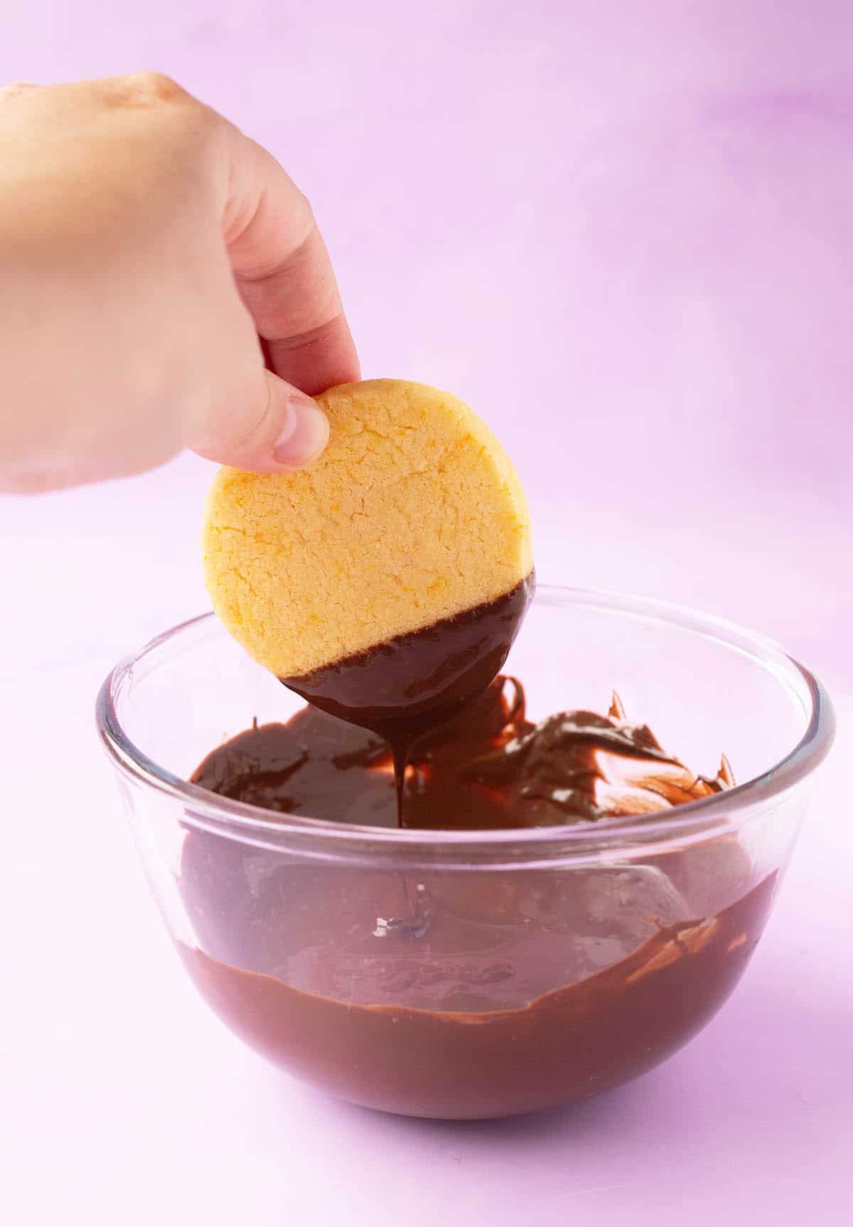 A hand dipped a shortbread cookie into a bowl of melted dark chocolate. 