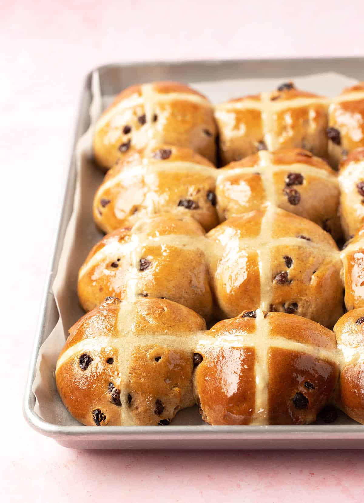 A pan filled with freshly glazed hot cross buns on a pink background.