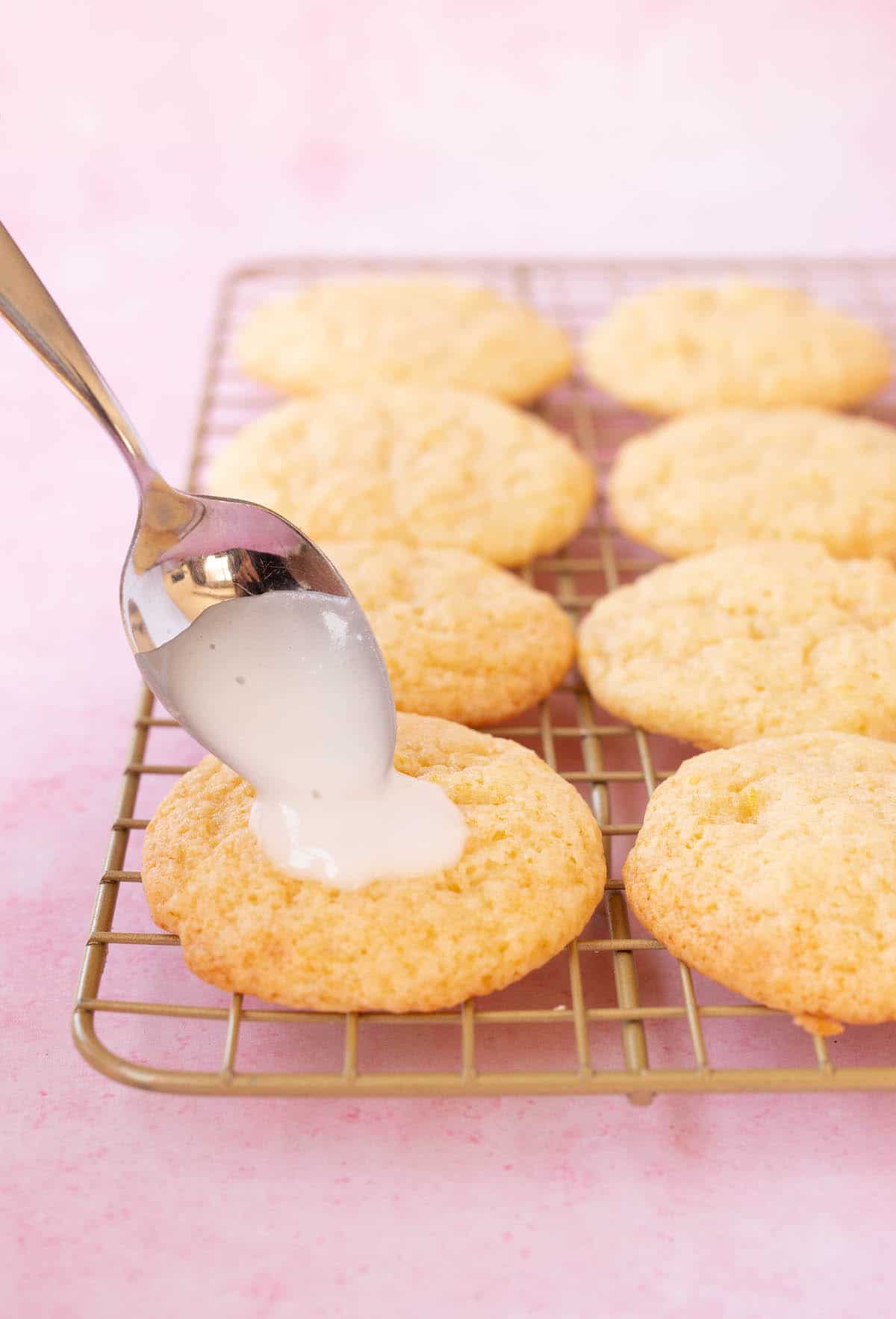 A gold wire rack with Lemon Ricotta Cookies cooling. A spoon adds lemon glaze icing.