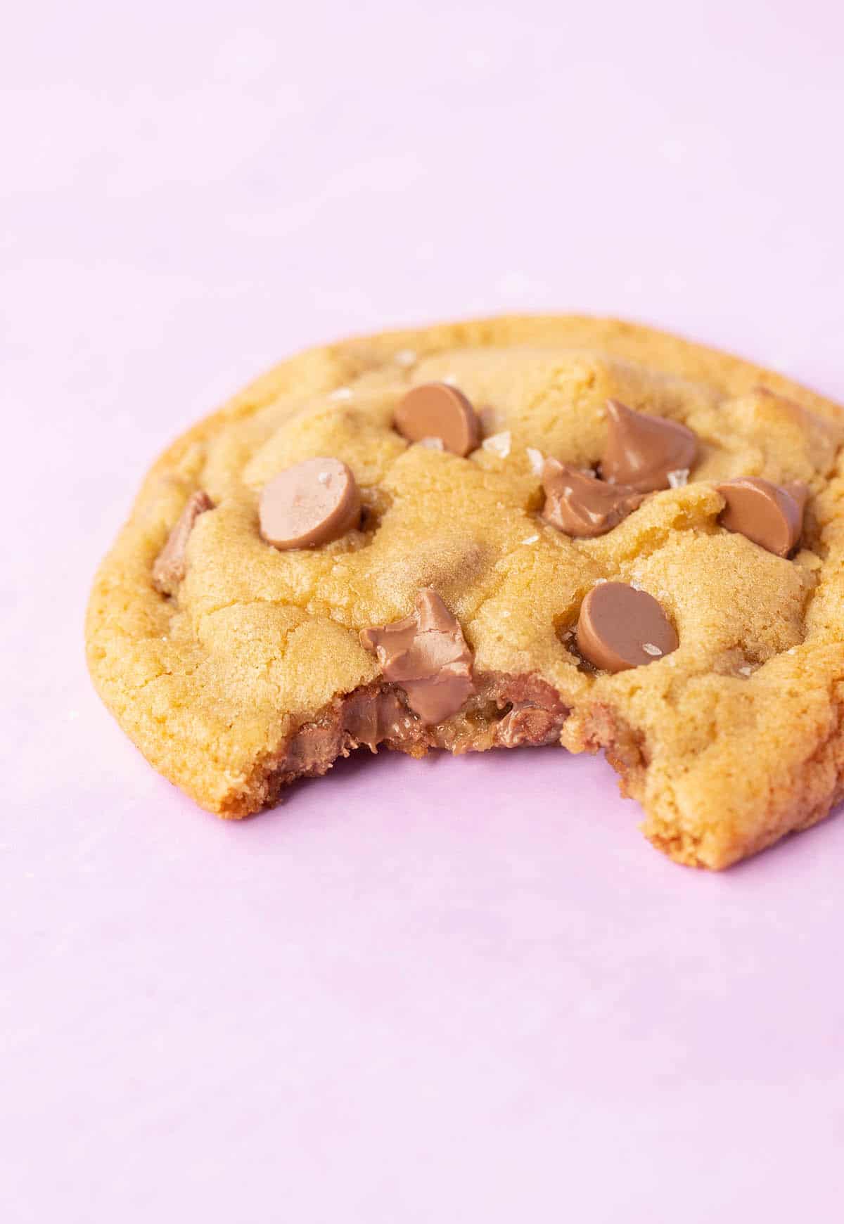 A single chocolate chip cookie with a bite taken out of it on a purple backdrop.