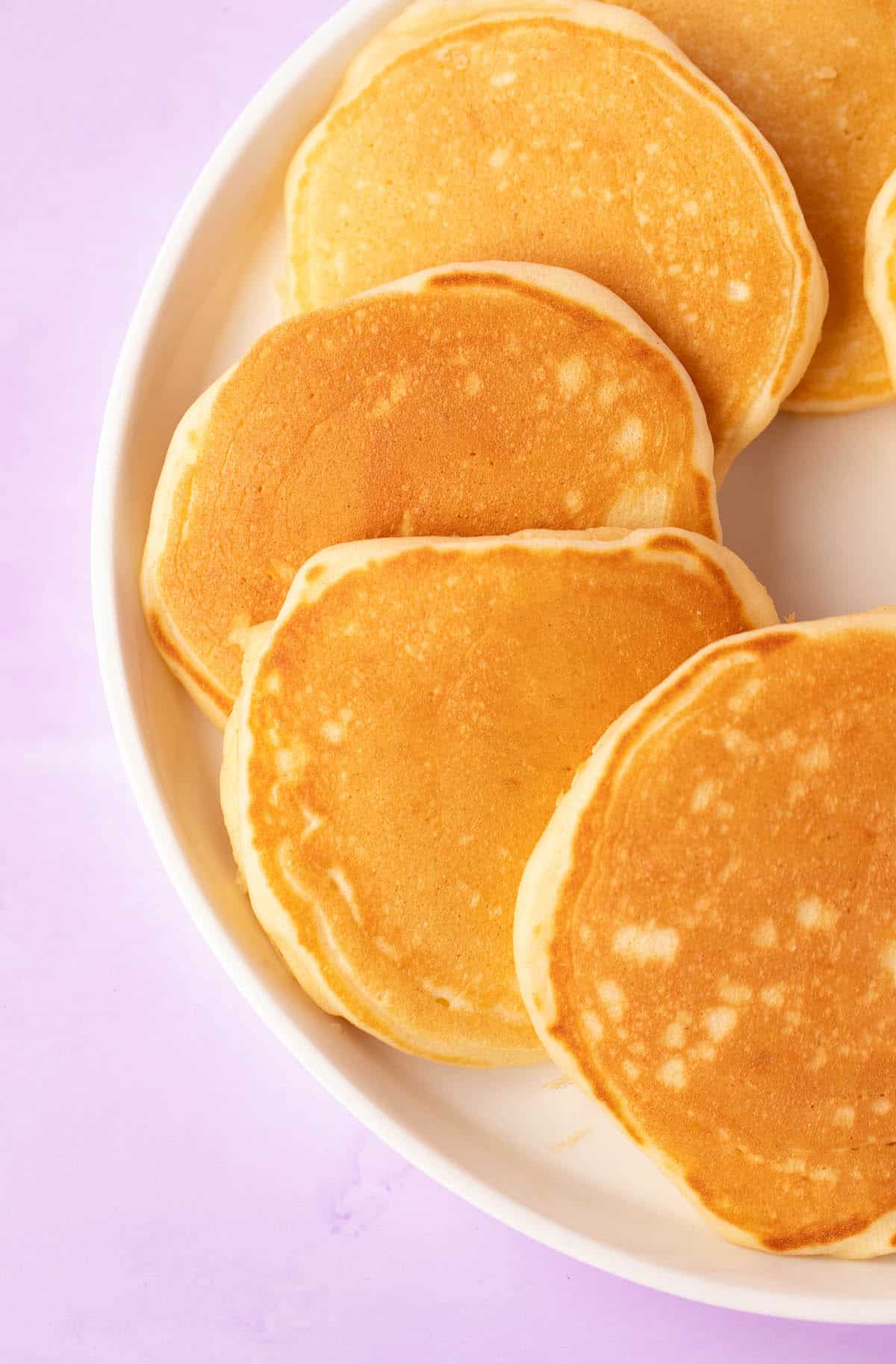Mini pancakes on a white plate with a purple background. 