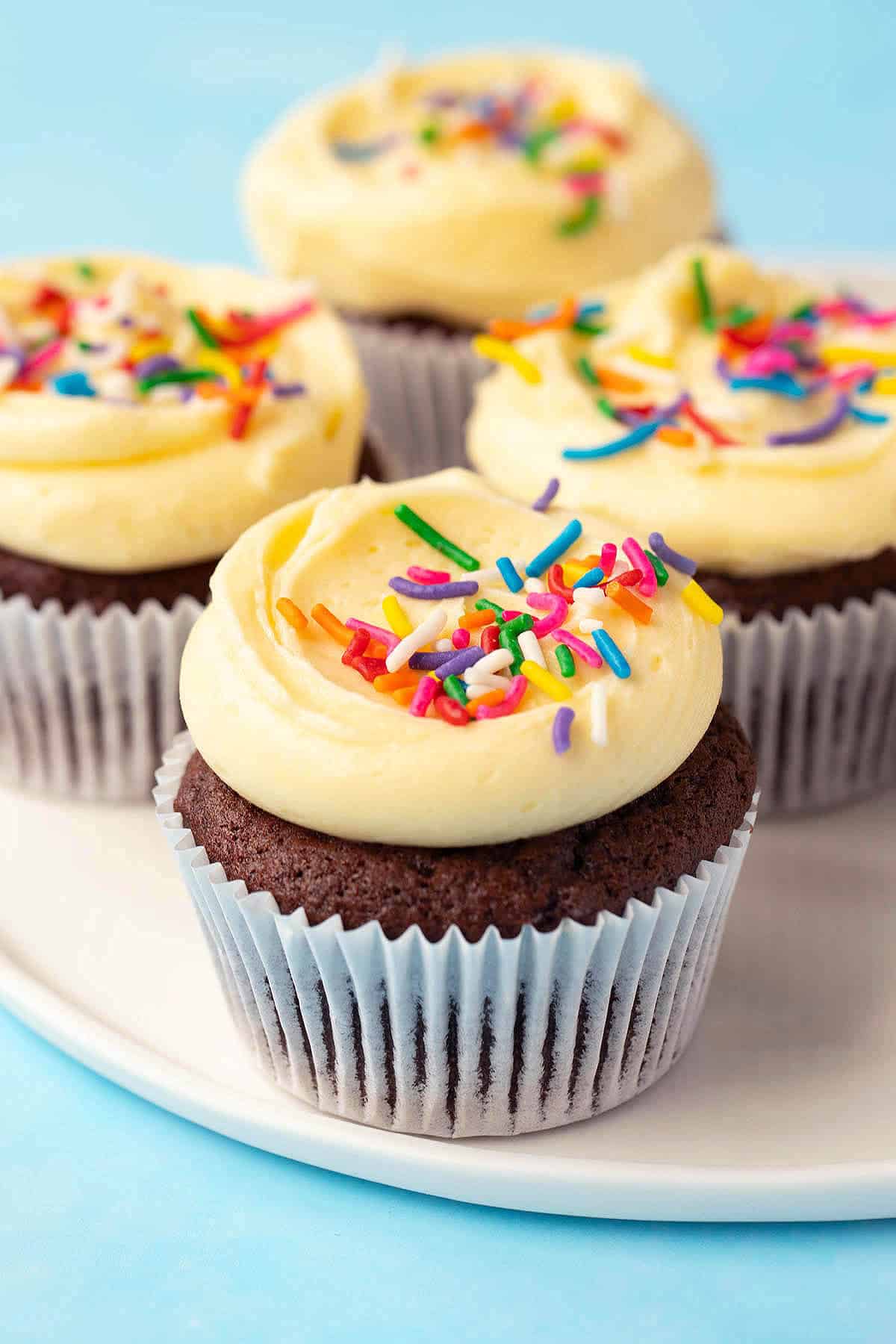A plate of chocolate cupcakes with yellow frosting and sprinkles on a white plate.