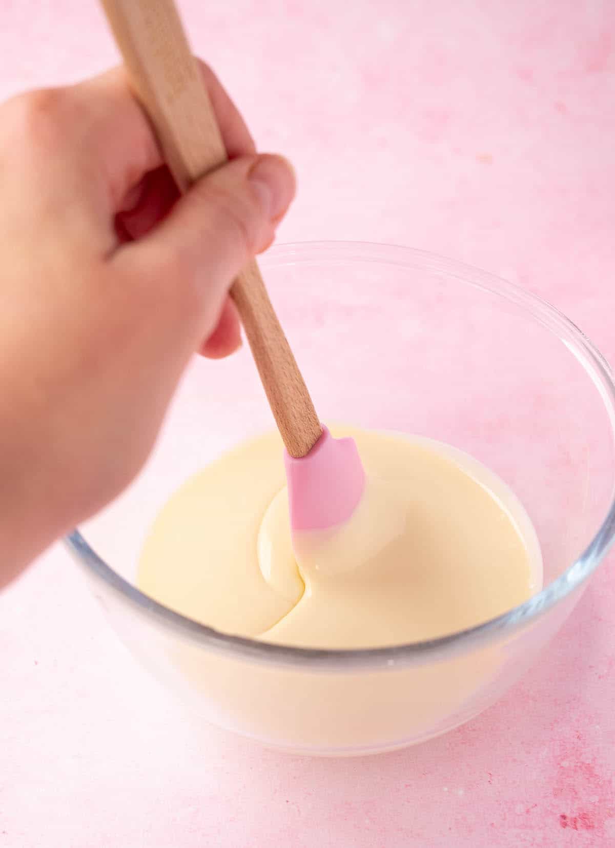 A glass bowl with melted white chocolate being stirred with a pink spatula.
