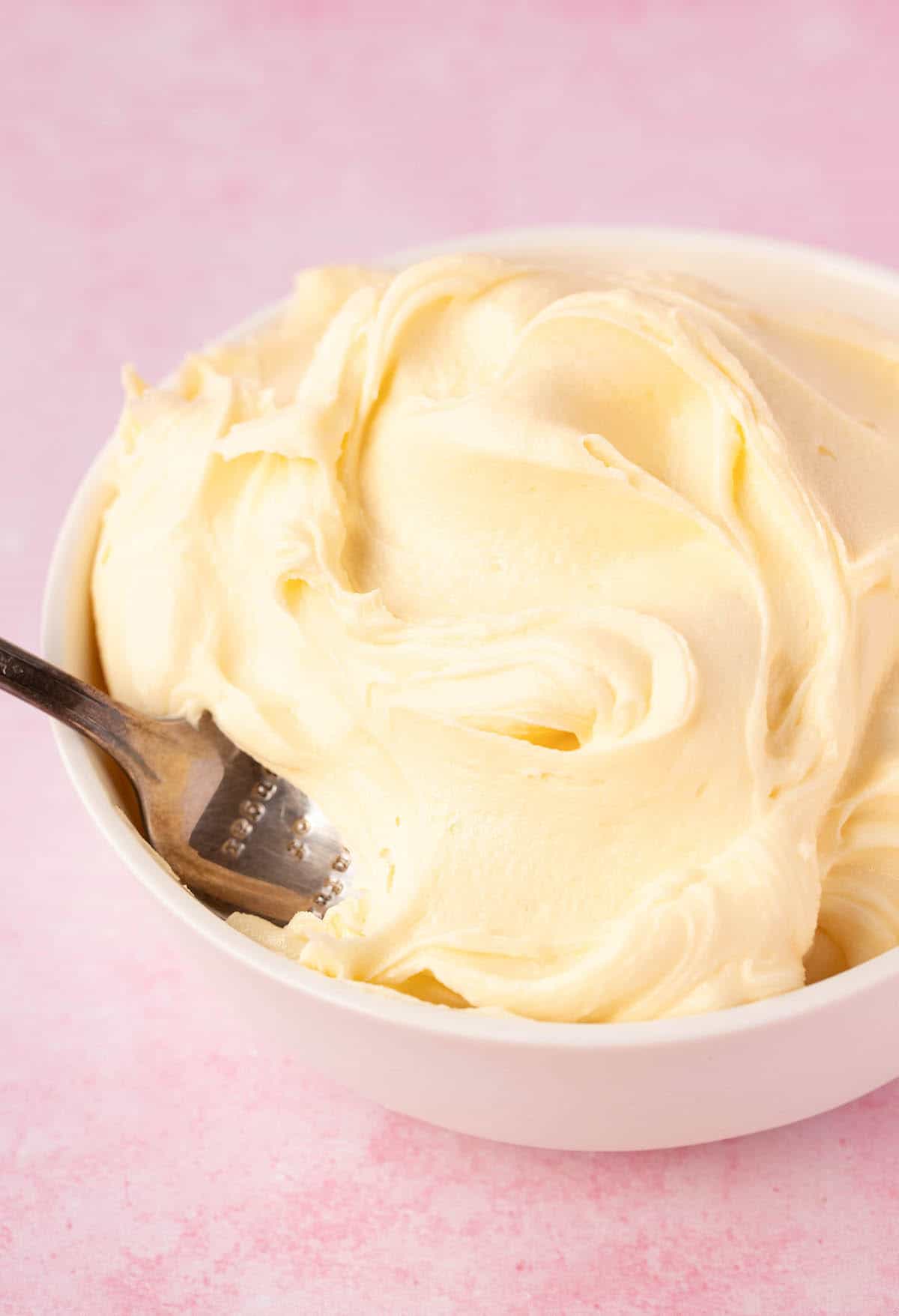 A white bowl filled with creamy white chocolate frosting on a pink background.