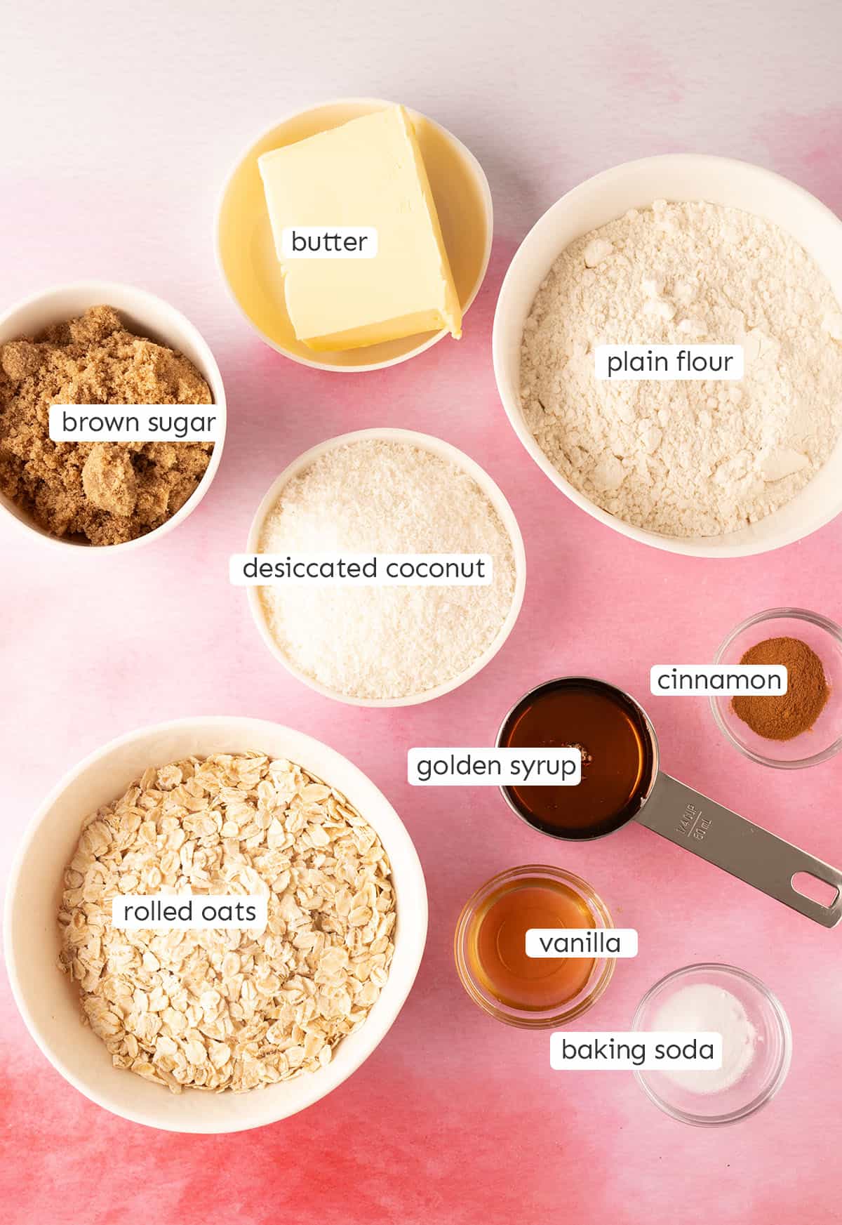 All the ingredients needed to make Anzac Biscuits from scratch on a pink backdrop.