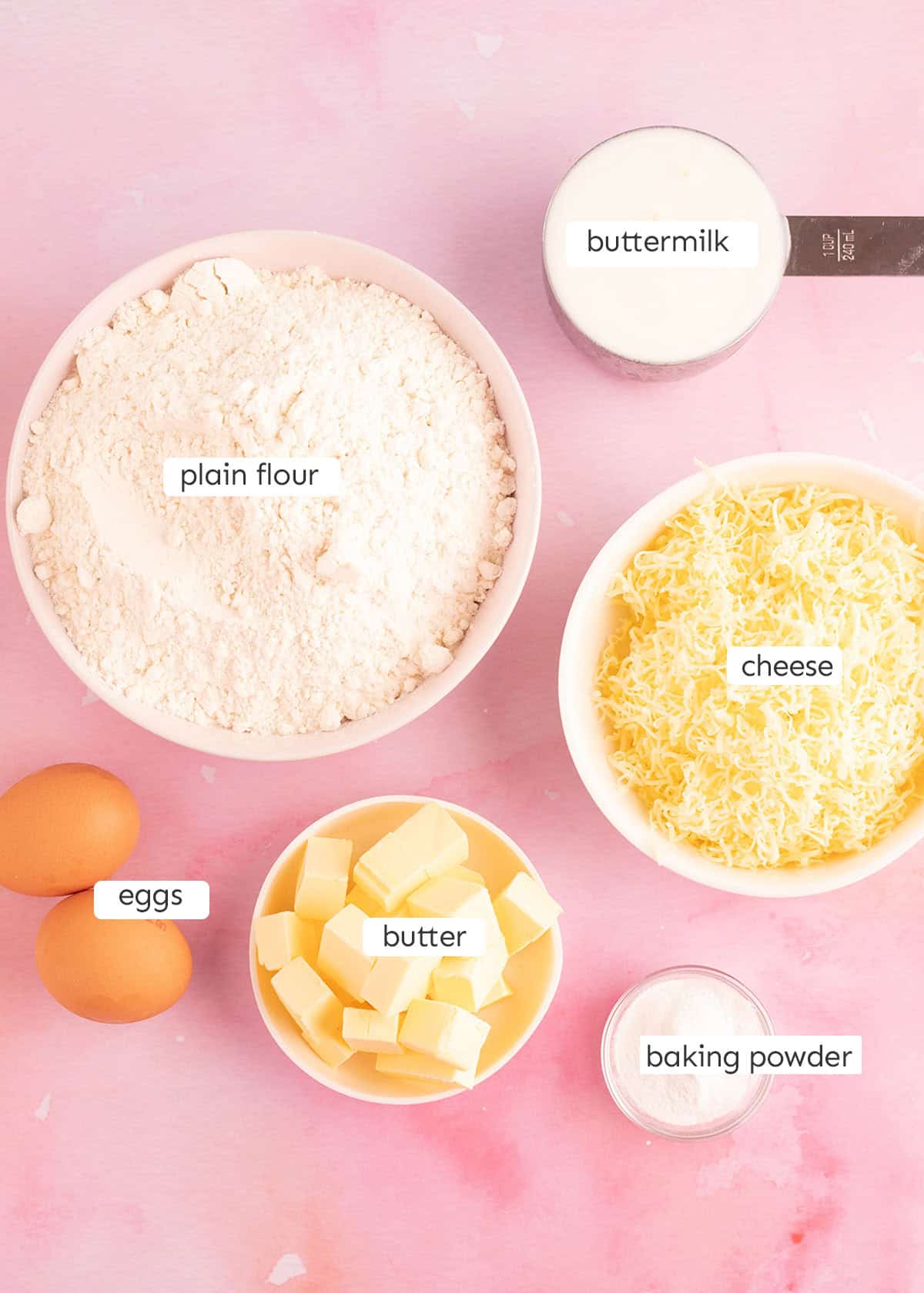 All the ingredients needed to make cheese scones from scratch on pink backdrop.